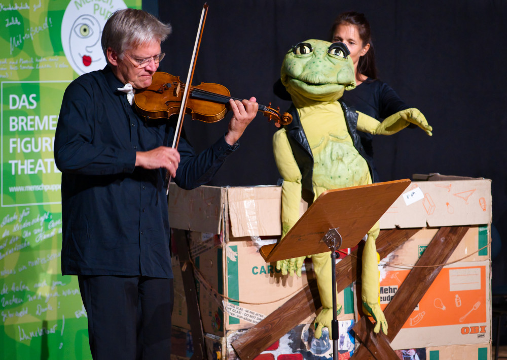 Violinist und Puppenspielerin mit lebensgroßer Froschpuppe